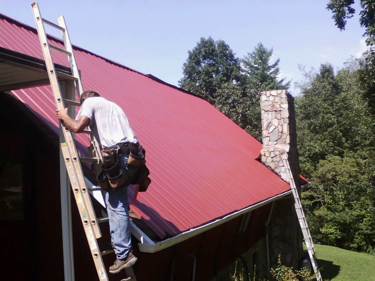 Roofer on a ladder beside a red metal roof and stone chimney — Soffit & Fascia Repair by Roofing Brothers in Strawberry Plains